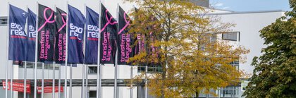 Eine Gruppe Fahnenmasten vor den Messehallen, mit den Logos von EXPO REAL sowie transform & beyond. Im Vordergrund ist ein See und Grünanlagen zu sehen.