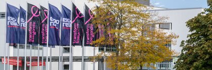 Eine Gruppe Fahnenmasten vor den Messehallen, mit den Logos von EXPO REAL sowie transform & beyond. Im Vordergrund ist ein See und Grünanlagen zu sehen.