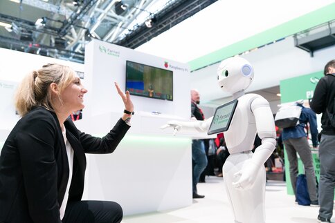 A woman gives a high-five to a humanoid robot at a trade fair stand.