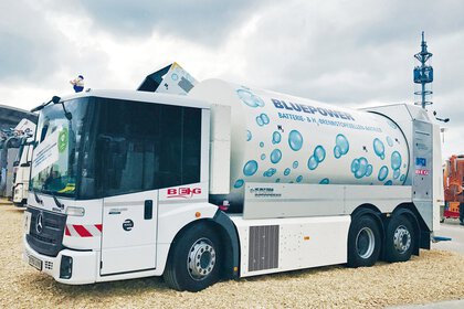 Weißes Demonstrationsfahrzeug mit großem Tankaufbau und der Aufschrift „BLUEPOWER – Batterie- & H₂-Brennstoffzellen-Antrieb“, dekoriert mit blauen Wasserstoffblasen, ausgestellt im Freien.