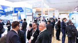 Numerous visitors to LOPEC stand in business attire in a corridor between different exhibition stands and talk to each other.