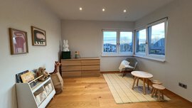 Minimalistisches Kinderzimmer mit Holzboden, Spielzeug, Büchern, einer Kommode und einem kleinen Tisch vor großen Fenstern.