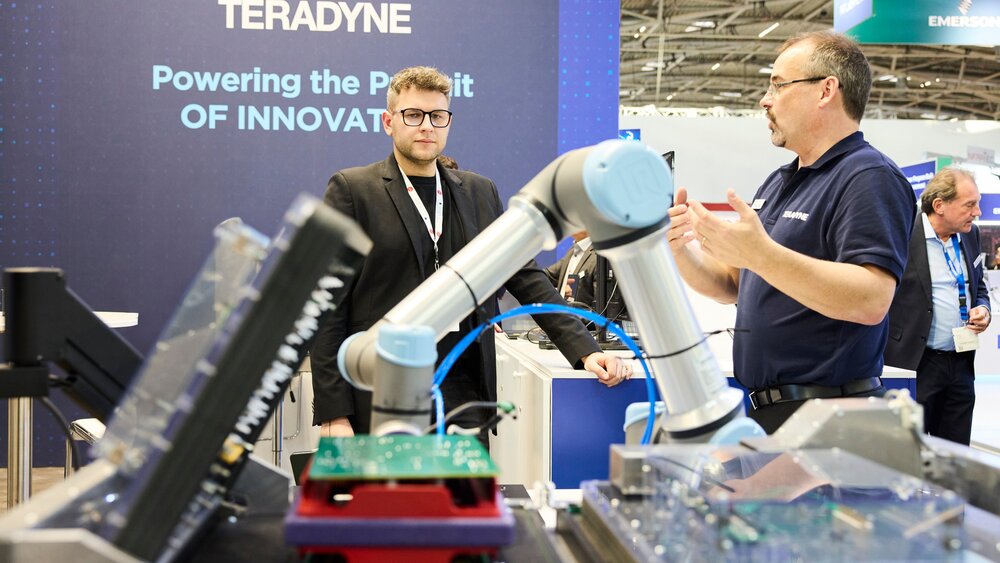 Zwei Männer stehen an einem Stand auf der productronica und betrachten einen einen silber-grau-blauen Roboterarm.
