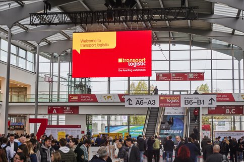 Entrance area of Messe München with numerous visitors, a large welcome sign for transport logistic hangs above. Signs point the way to Halls A6-A3 and B6-B1.