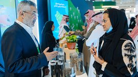  Several people are standing at a long counter at IFAT Saudi Arabia, discussing the displays presented on it.
