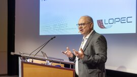 Ein Mann im grauen Anzug und mit Brille steht an einem Podium vor einer Präsentationsleinwand und hält einen Vortrag.