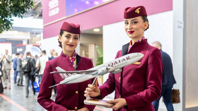 Two young dark-haired women in burgundy stewardess uniforms and small hats hold a model aeroplane with the inscription Qatar. The exhibition hall is blurred in the background.