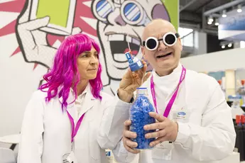 A woman in a pink wig and a man with round lab goggles, both in lab coats, present a product - a jar filled with blue pearls.
