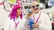 Frau mit pinker Perücke und ein Mann mit runder Laborbrille, beide im Laborkittel, präsentieren ein Produkt - ein Gefäß gefüllt mir blauen Perlen.