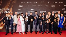 Sixteen smiling people in festive attire stand together in front of a photo wall of the INHORGENTA AWARD 2026, holding their trophies in their hands.
