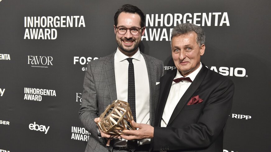 Two men smile and proudly hold the trophy in the “Gemstone Design of the Year” category in front of a logo wall at the AWARD Gala.
