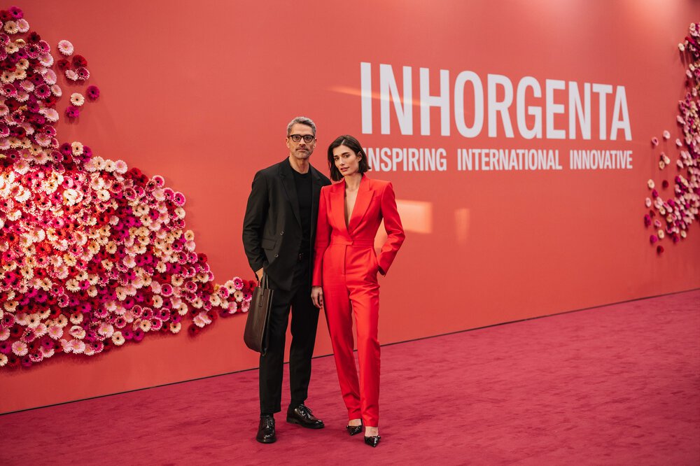 Ein Mann und eine Frau in Business-Kleidung stehen auf dem roten Teppich der INHORGENTA vor einer roten Wand mit Blumendokoration.