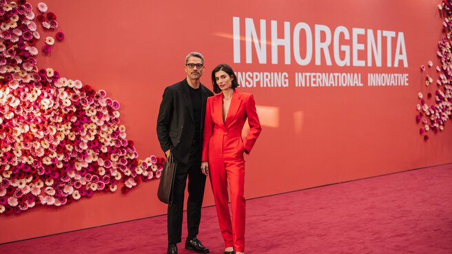 A man and a woman in business attire stand on the INHORGENTA red carpet in front of a red wall decorated with flowers.