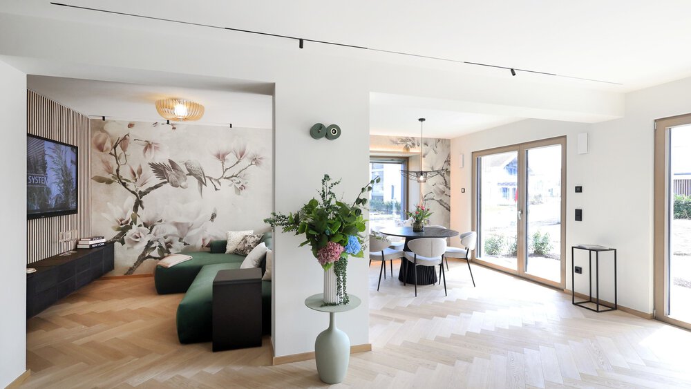 Moderner möblierter Wohn- und Essbereich mit großen Fenstern, floraler Wandtapete und hellen Holzböden.