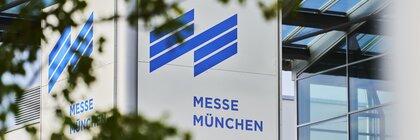 Nahansicht des Messe München Logos über dem Schild „Eingang West“ vor der Eingangshalle und umrahmt von grünen Blättern.