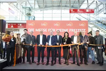 A group of people stand on a stage at the opening ceremony of a trade fair and look into the camera. They are all holding the opening ribbon that is to be cut.