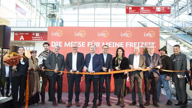 Eine Gruppe von Menschen steht auf einer Bühne bei einer Eröffnungsfeier einer Messe und schaut in die Kamera. Alle halten das Eröffnungsband in der Hand, dass zerschnitten werden soll.