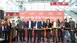 A group of people stand on a stage at the opening ceremony of a trade fair and look into the camera. They are all holding the opening ribbon that is to be cut.