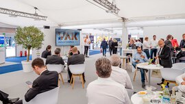 Ein Konferenzraum mit sitzenden und stehenden Menschen, zwischen denen auf einem Bildschirm der VAK Innovationspreis 2022 präsentiert wird.