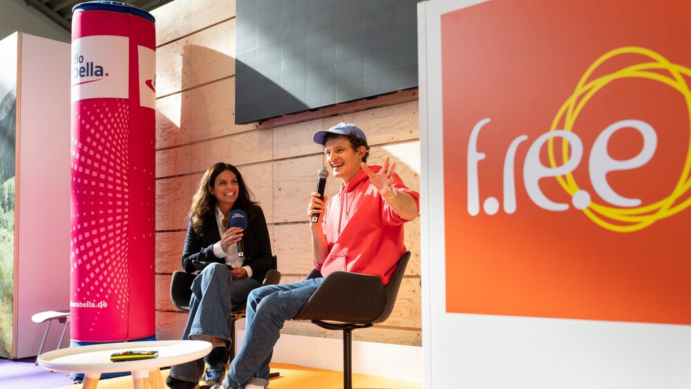 A presenter and a guest are sitting on stage talking, holding microphones in their hands. Next to them, the logo of the leisure fair can be seen on a large board.
