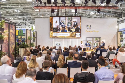 The audience is watching a panel discussion on the IFAT Munich Orange Stage, with the speakers on stage displayed on a large screen.