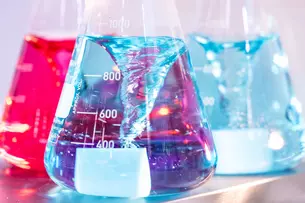 Erlenmeyer flasks with red, blue and light blue liquids, which are partially filled and stand in front of a blurred background.