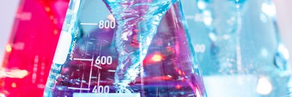 Erlenmeyer flasks with red, blue and light blue liquids, which are partially filled and stand in front of a blurred background.
