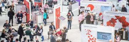 Ein großer Ausstellerstand auf der analytica mit einigen Stellwänden und vielen Personen. Einige führen Gespräche, auch an Tischen sitzend.