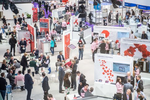 Ein großer Ausstellerstand auf der analytica mit einigen Stellwänden und vielen Personen. Einige führen Gespräche, auch an Tischen sitzend.