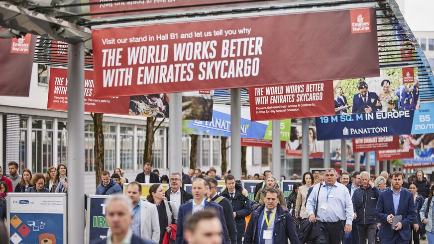Zahlreiche Besucher der transport logistic laufen unter einem Vordach entlang, an dem viele bunte Werbeplakate von teilnehmenden Ausstellern hängen.
