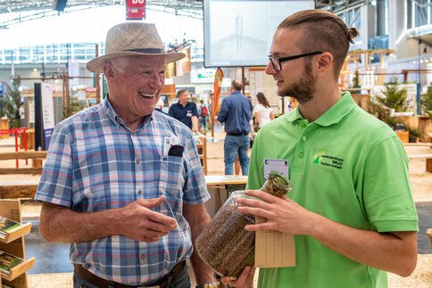 A visitor in a chequered shirt and an INTERFORST exhibitor in a green shirt discuss a bottle of seed in the exhibition hall.