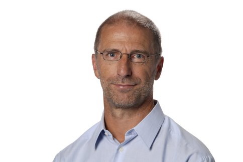 Ein Mann mit sehr kurzen Haaren, Bart und Brille. Er trägt ein hellblaues Hemd und lächelt.