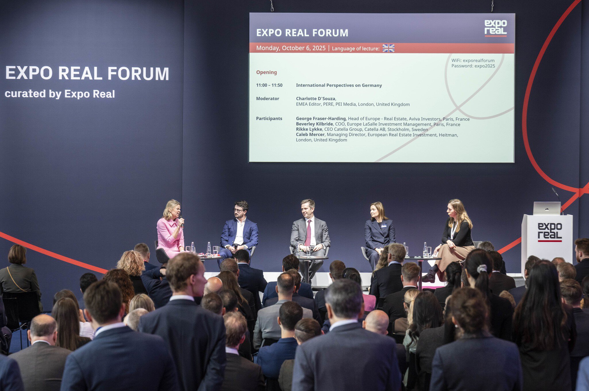 Eine Podiumsdiskussion beim EXPO REAL Forum in München. Fünf Expertinnen und Experten sitzen auf der Bühne und sprechen über internationale Perspektiven auf den deutschen Immobilienmarkt. Die Moderatorin im pinkfarbenen Kleid hält ein Mikrofon, während das Publikum aufmerksam zuhört. Auf der Leinwand im Hintergrund sind Programmdetails mit den Namen der Teilnehmenden und dem Veranstaltungstitel „EXPO REAL Forum – Monday, October 6, 2025“ zu sehen.