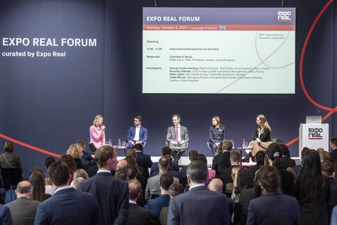 A panel discussion at the EXPO REAL Forum in Munich. Five experts are seated on stage discussing international perspectives on the German real estate market. The moderator in a pink dress is speaking into a microphone while the audience listens attentively. A large screen behind them displays the program details, including participant names and the event title “EXPO REAL Forum – Monday, October 6, 2025.”