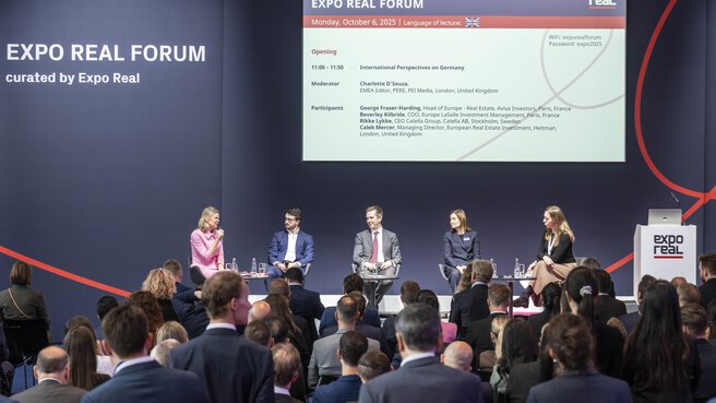 Eine Podiumsdiskussion beim EXPO REAL Forum in München. Fünf Expertinnen und Experten sitzen auf der Bühne und sprechen über internationale Perspektiven auf den deutschen Immobilienmarkt. Die Moderatorin im pinkfarbenen Kleid hält ein Mikrofon, während das Publikum aufmerksam zuhört. Auf der Leinwand im Hintergrund sind Programmdetails mit den Namen der Teilnehmenden und dem Veranstaltungstitel „EXPO REAL Forum – Monday, October 6, 2025“ zu sehen.