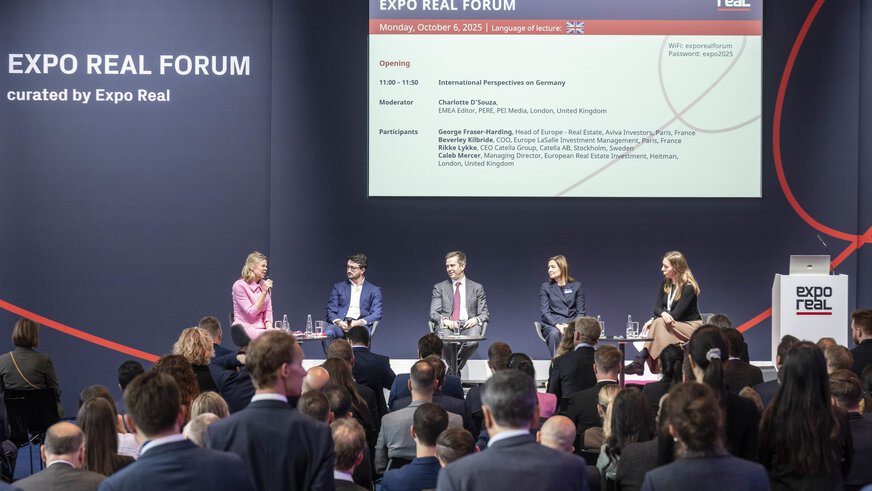 A panel discussion at the EXPO REAL Forum in Munich. Five experts are seated on stage discussing international perspectives on the German real estate market. The moderator in a pink dress is speaking into a microphone while the audience listens attentively. A large screen behind them displays the program details, including participant names and the event title “EXPO REAL Forum – Monday, October 6, 2025.”