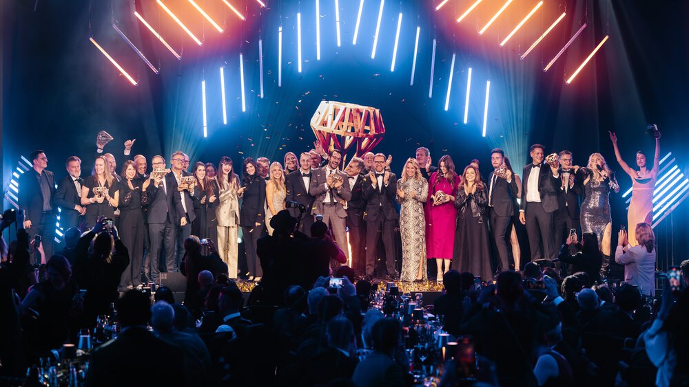 Eine große Gruppe von Menschen in formeller Kleidung steht auf einer Bühne und feiert unter hellem Neonlicht vor dem Publikum.