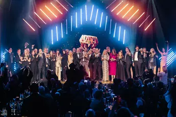 A large group of people in formal dress stand on a stage and celebrate in front of the audience under bright neon lights.