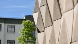 Close-up of a modern building facade with geometric panels, trees, and another building in the background.