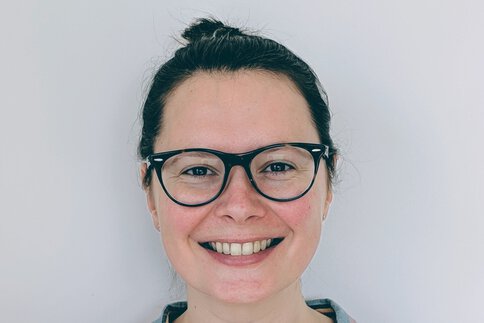 Eine lächelnde Frau mit schwarzer Brille und zurückgebundenen schwarzen Haaren. Sie trägt ein bunt gestreiftes Hemd.