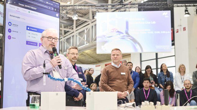 a man in a shirt stands in front of a laboratory device with a microphone and explains something