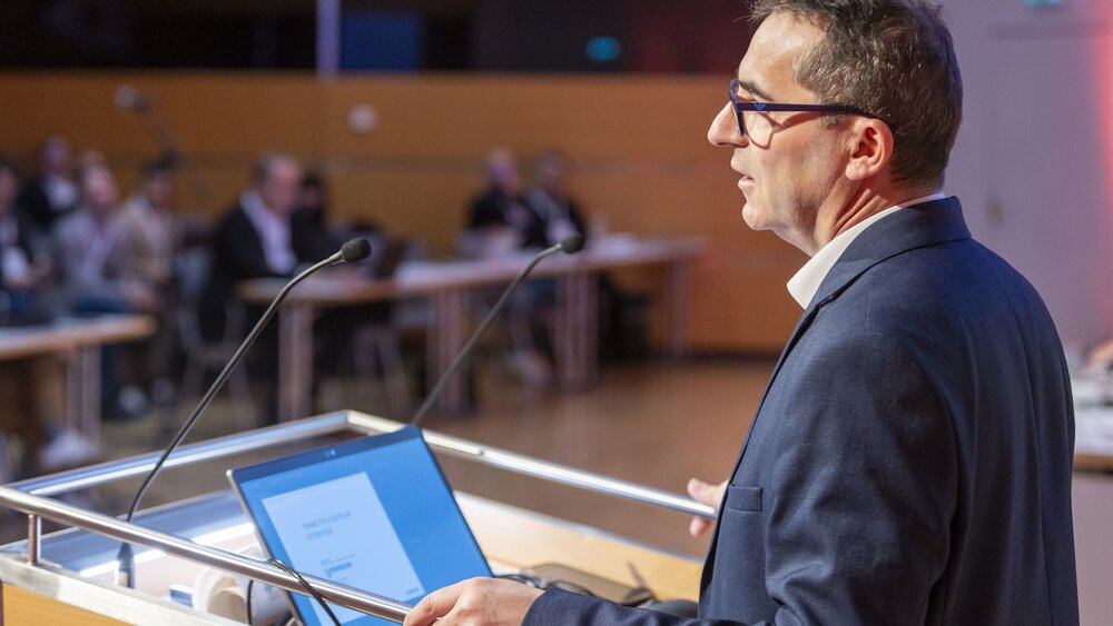 Ein Mann im Anzug und mit Brille steht an einem Podium vor einem aufgeklappten Laptop und hält einem Publikum einen Vortrag.
