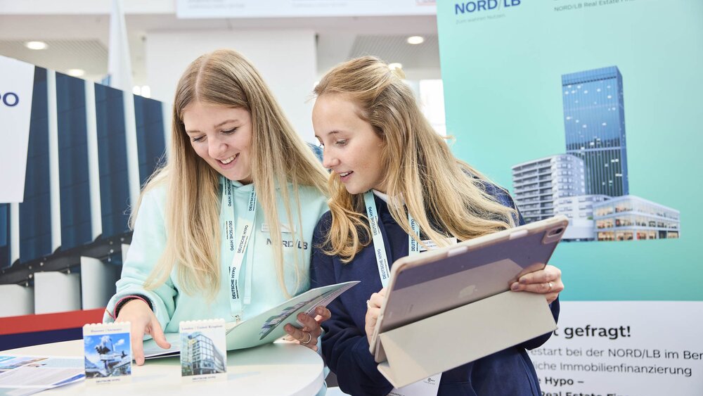 Zwei junge Frauen stehen an einem Messestand und schauen gemeinsam auf Informationsmaterial. Eine hält ein Tablet, die andere blättert durch eine Broschüre. Beide tragen NORD/LB-Lanyards und lächeln, während im Hintergrund ein Roll-up mit Gebäudedruck zu sehen ist.