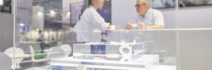 Eine Vitrine mit Schaustücken auf einem Messestand, im Hintergrund sind unscharf zwei Personen zu sehen, die sich an einem Tresen unterhalten.