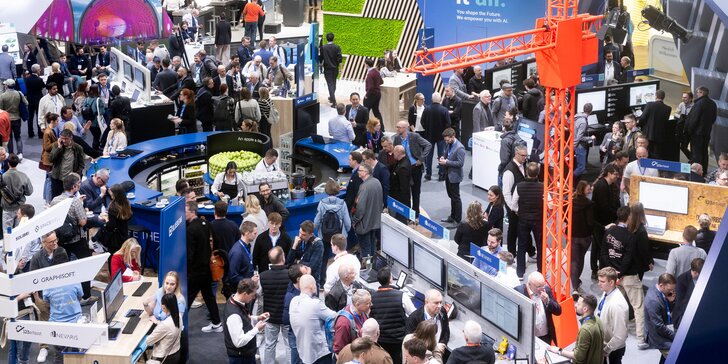 Densely packed trade fair hall with numerous booths for digital or technological solutions, where visitors interact, watch presentations, and work with displays.