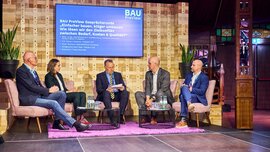Fünf Personen sitzen in Sesseln auf einer Bühne und diskutieren in einer moderierten Gesprächsrunde der Veranstaltung „BAU PreView“, im Hintergrund zeigt eine Leinwand den Titel „Einfacher bauen, klüger umbauen: Wie lösen wir den Zielkonflikt zwischen Bedarf, Kosten & Qualität?“.