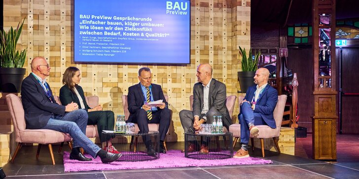 Five people sit in armchairs on a stage and take part in a moderated discussion round at the “BAU PreView” event, with a screen in the background showing the title “Building more simply, converting more intelligently: How do we resolve the conflict between demand, costs & quality?”.