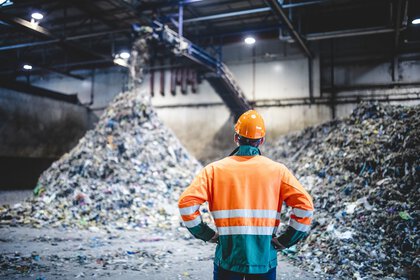 Ein Mann mit Helm und Arbeitsbekleidung steht in einer Halle, in der mit einem Förderband Müll sortiert wird.