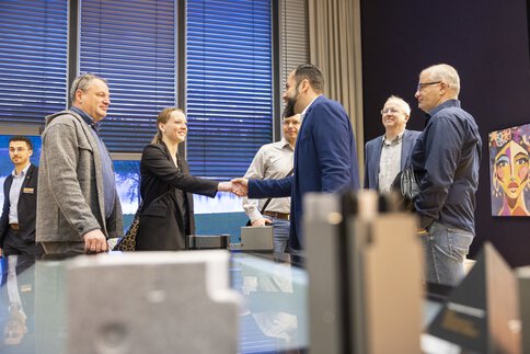 Sechs Personen in Businesskleidung stehen zusammen in einem Raum mit hohen Fenstern und Kunst an der Wand. Zwei der Personen schütteln einander die Hand.