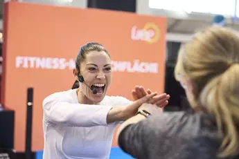 In the fitness participation area at f.re.e 2025, a trainer with a headset motivates a visitor to do sports exercises.